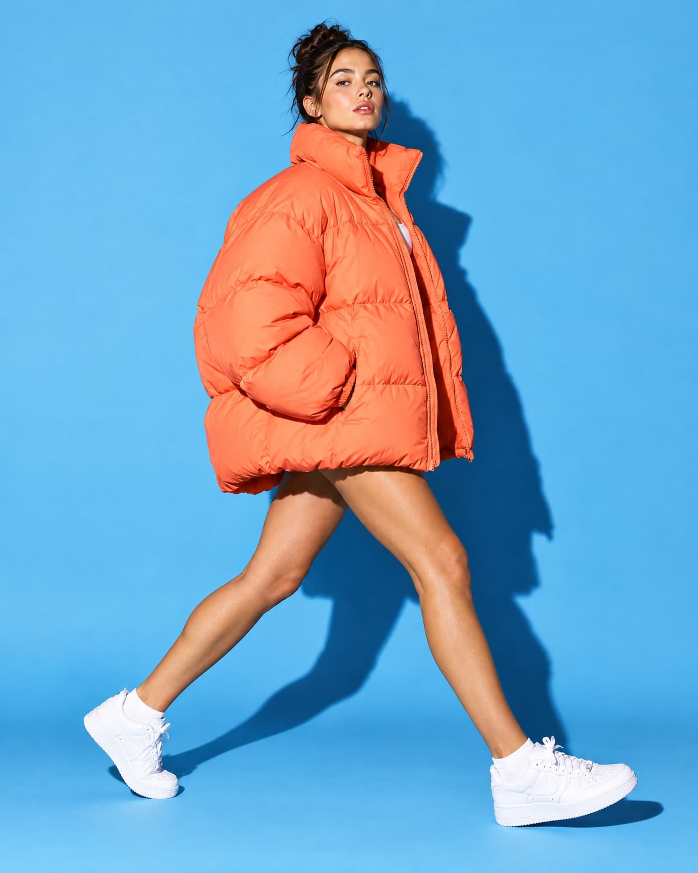 Fashion ad creative: a model in a coral puffer jacket striding across a sky blue backdrop