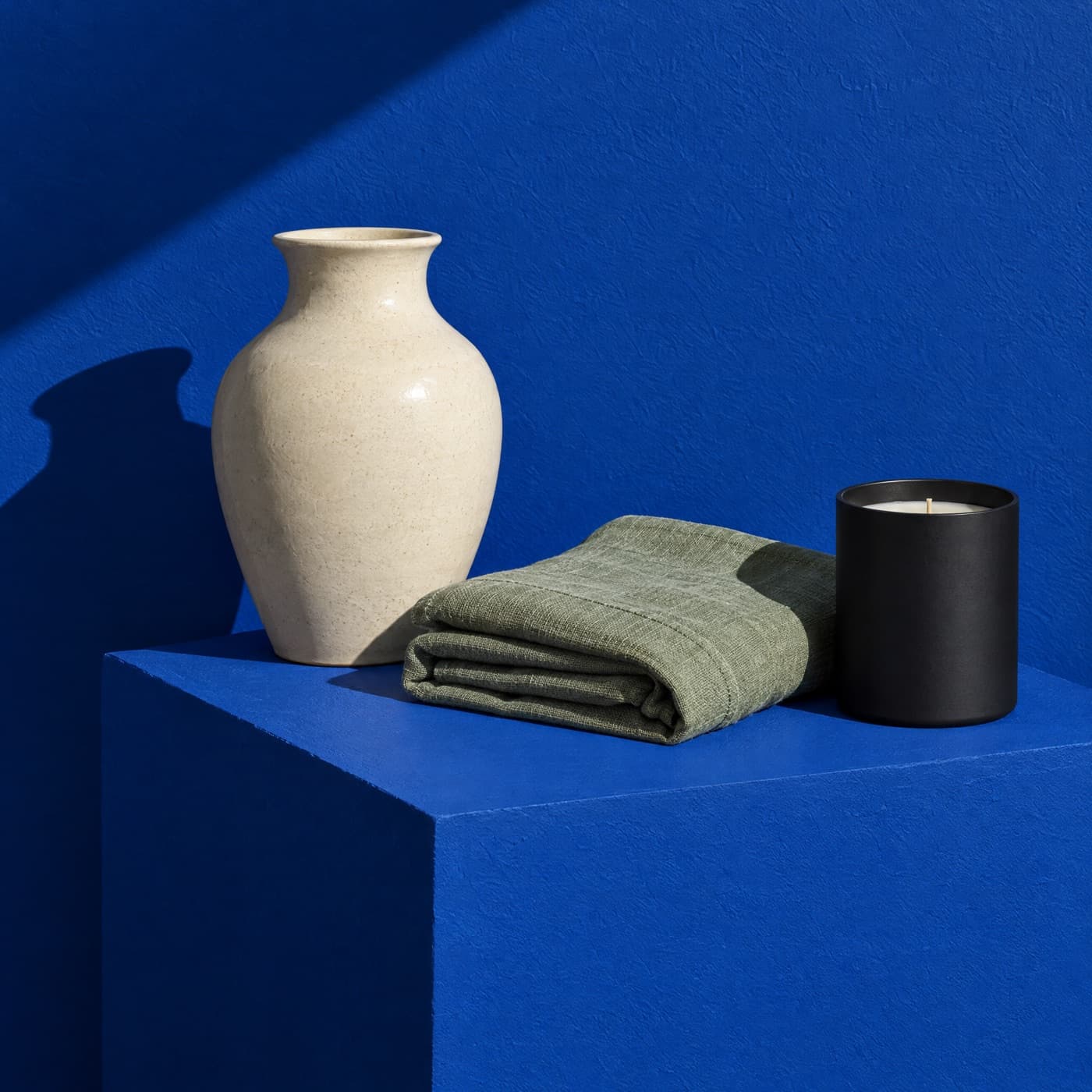 A cream vase, folded sage linen and a black candle on a cobalt blue block